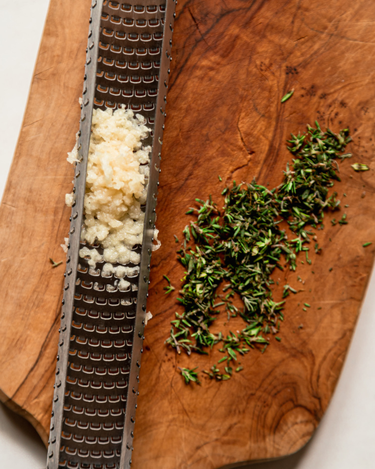 An overhead shot shows finely grated garlic in a Microplane and minced fresh thyme on a wood cutting board.