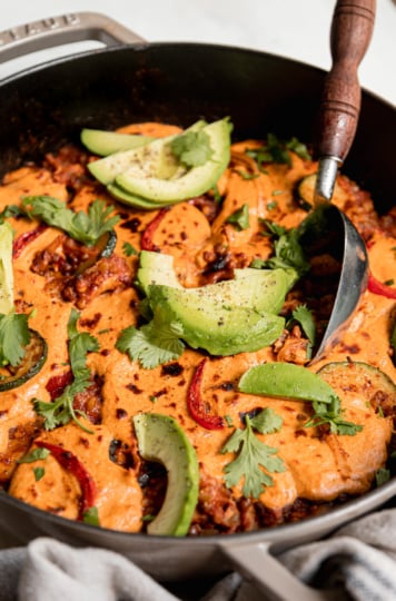 A 3/4 angle shot of lazy chopped vegan enchiladas in a skillet. A sunflower "cheese" topping blankets the top and is browned in spots. The skillet is garnished with sliced avocado and chopped cilantro.