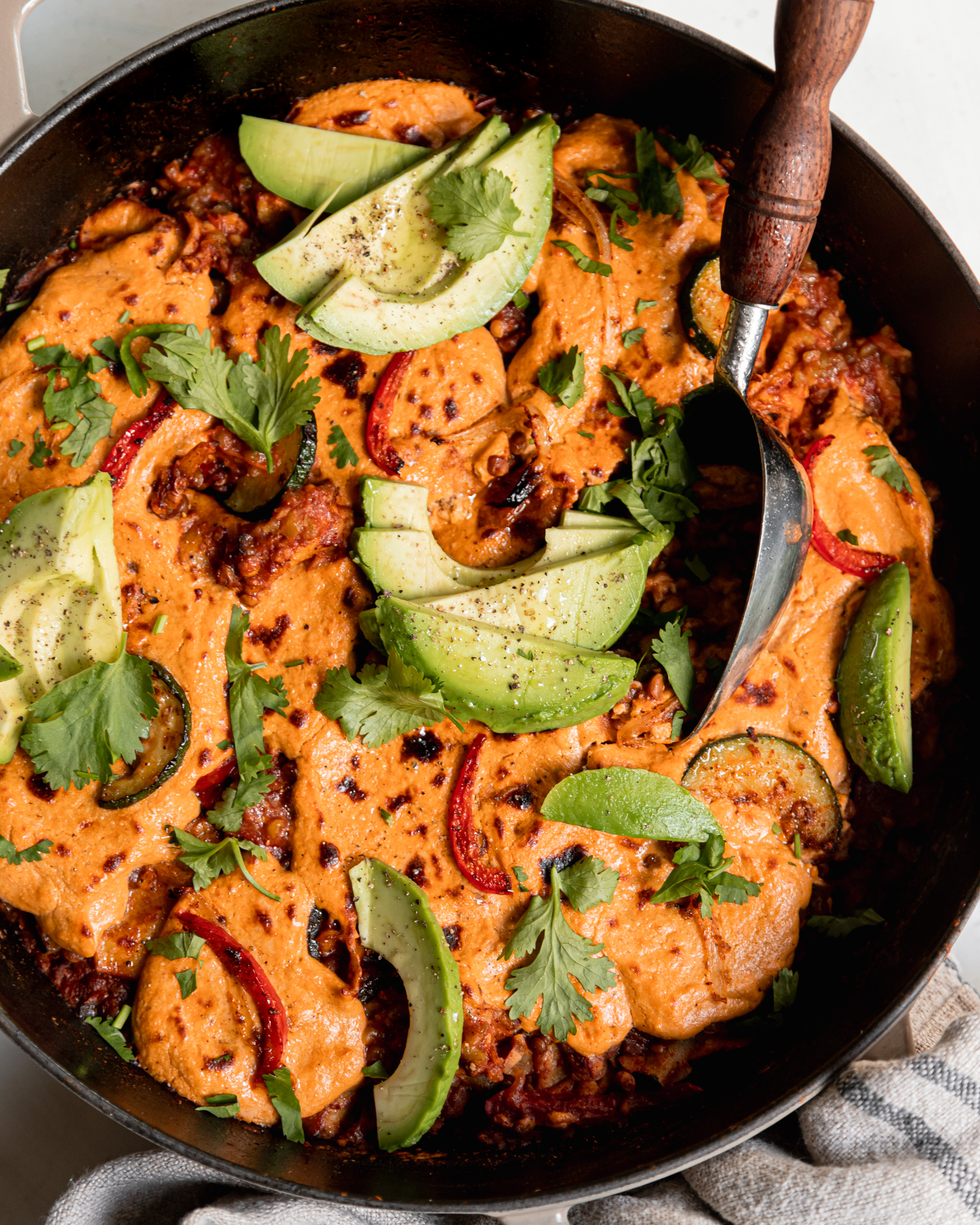 An overhead shot of lazy chopped vegan enchiladas in a skillet. A sunflower "cheese" topping blankets the top and is browned in spots. The skillet is garnished with sliced avocado and chopped cilantro.