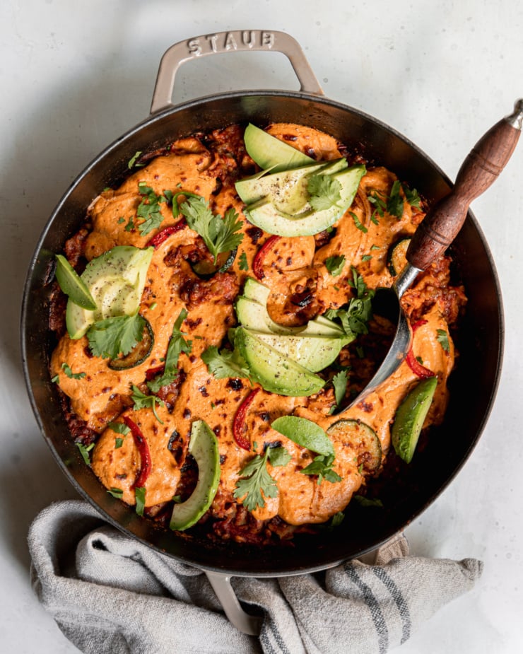 An overhead shot of lazy chopped vegan enchiladas in a skillet. A sunflower "cheese" topping blankets the top and is browned in spots. The skillet is garnished with sliced avocado and chopped cilantro.