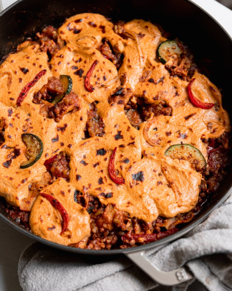 An overhead shot of lazy chopped vegan enchiladas in a skillet. A sunflower "cheese" topping blankets the top and is browned in spots. You can see the red enchilada sauce and lentil filling poking out in spots.