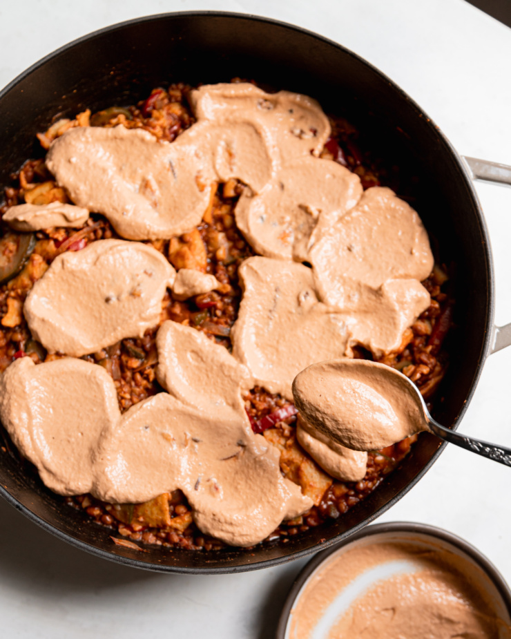 A spoon is applying a sunflower seed based "cheese" topping to a vegan enchilada skillet that features lentils and vegetables as the "filling."