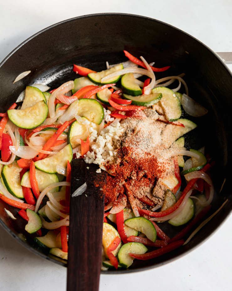 An overhead shot shows a braiser filled with sautรฉed peppers, zucchini, and onions with garlic and spices freshly added.