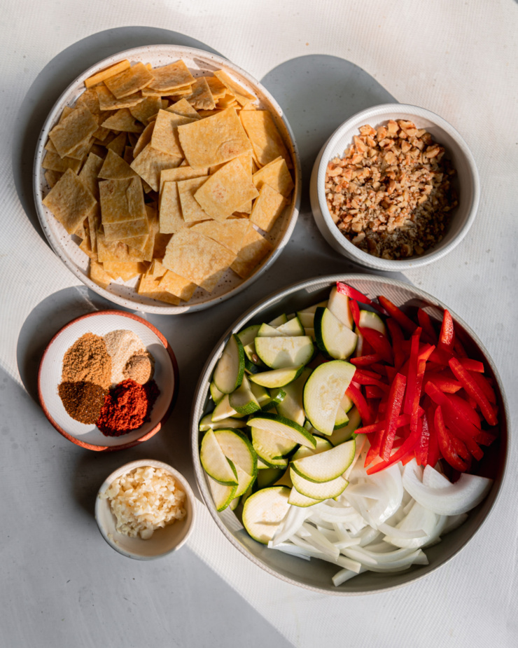 An overhead shot shows prepped ingredients in bowls: sliced vegetables, chopped tortillas, measured spices, finely chopped walnuts, and minced garlic.