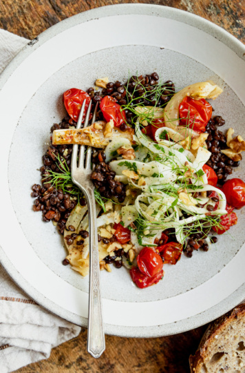 An overhead shot shows a wide serving bowl containing red wine French lentils, confit fennel slices and cherry tomatoes, a fresh shaved fennel salad on top, and a piece of whole grain bread to the side.