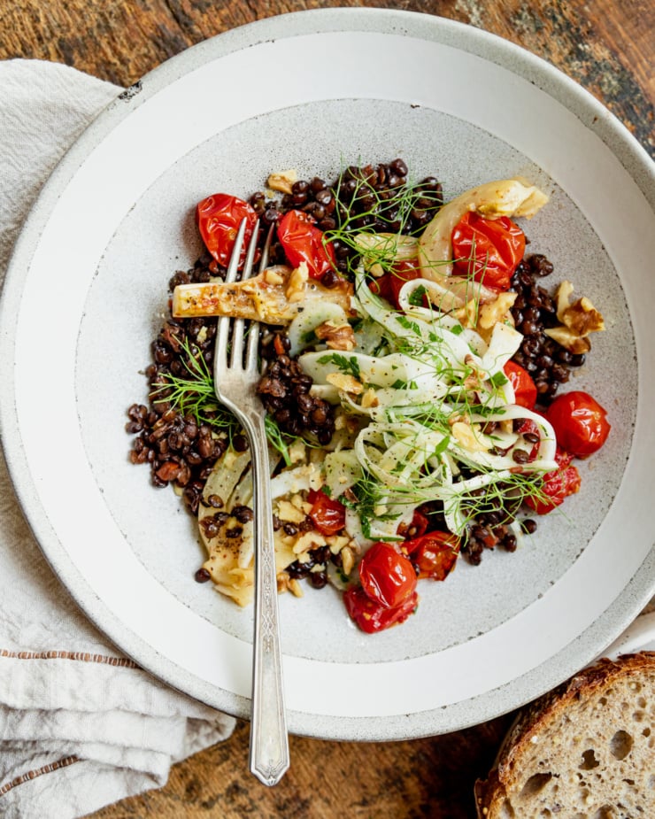 An overhead shot shows a wide serving bowl containing red wine French lentils, confit fennel slices and cherry tomatoes, a fresh shaved fennel salad on top, and a piece of whole grain bread to the side.