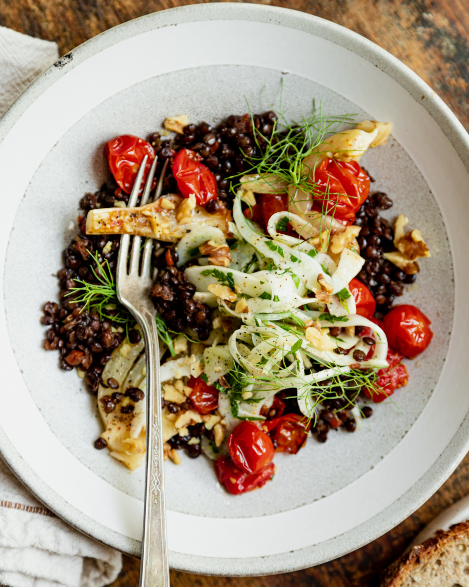 An up close, overhead shot shows a wide serving bowl containing red wine French lentils, confit fennel slices and cherry tomatoes, a fresh shaved fennel salad on top, and a piece of whole grain bread to the side.