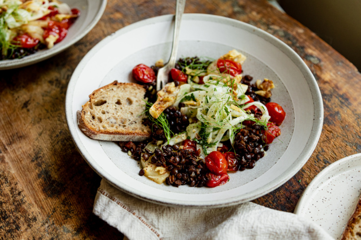 A 3/4 angle image shows a wide serving bowl containing red wine French lentils, confit fennel slices and cherry tomatoes, a fresh shaved fennel salad on top, and a piece of whole grain bread to the side.