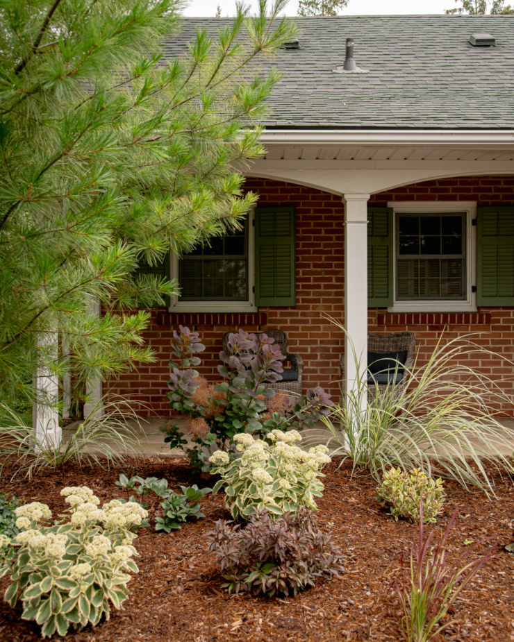 A head-on shot shows the front of a home with a garden in front of a porch area. The garden is covered in mulch and there is a pine tree swooping in from the left.