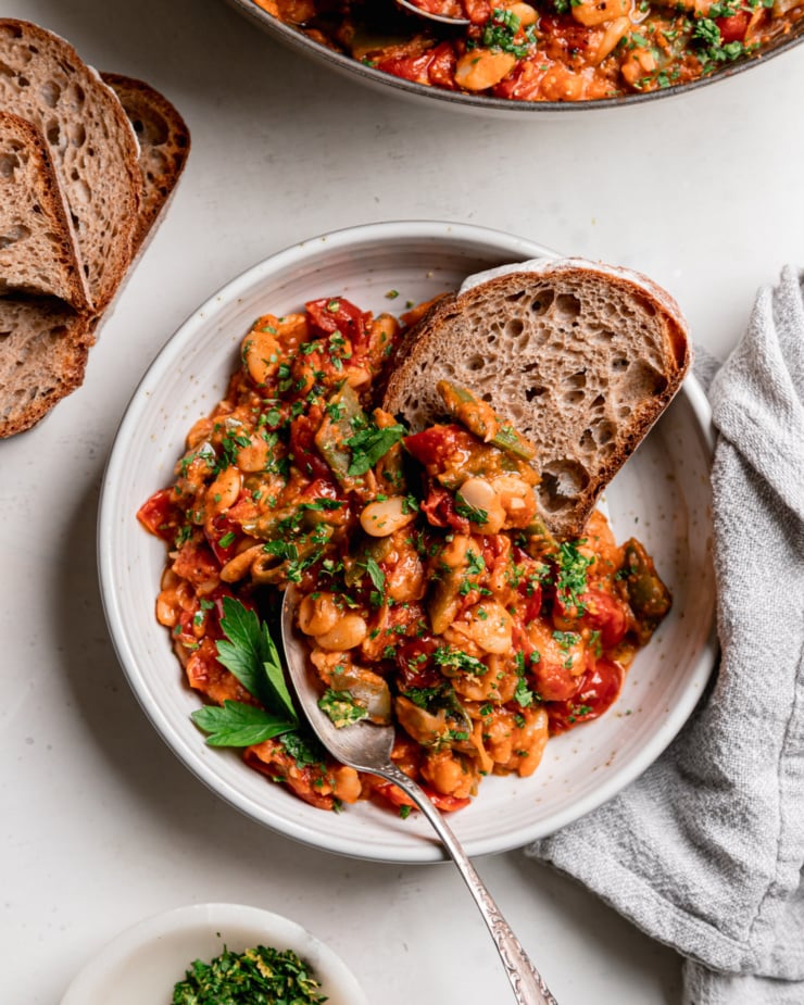 An overhead shot shows a single serving of cherry tomato-sauced butter beans and green romano beans in a shallow bowl. Some whole wheat sourdough bread is sticking out of the heap of beans and a spoon is in the bowl as well.