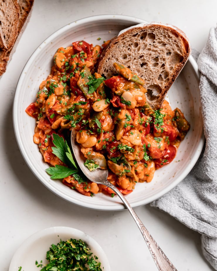 An overhead shot shows a single serving of cherry tomato-sauced butter beans and green romano beans in a shallow bowl. Some whole wheat sourdough bread is sticking out of the heap of beans and a spoon is in the bowl as well.
