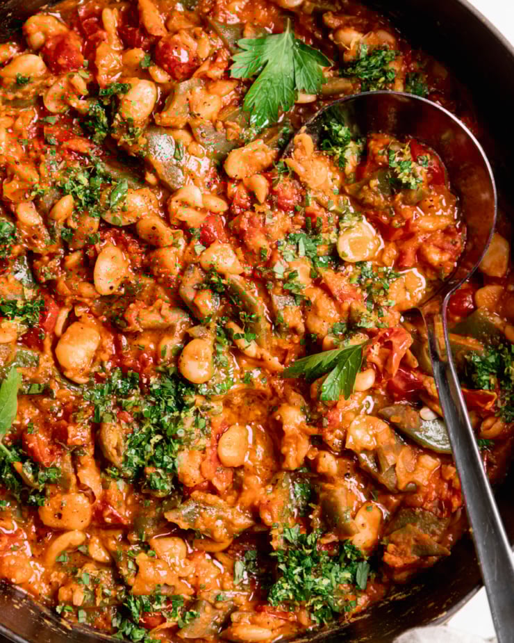 An up close, overhead shot shows a saucy butter bean skillet with a cherry tomato sauce and parsley on top. A ladle is sticking out of the pot.