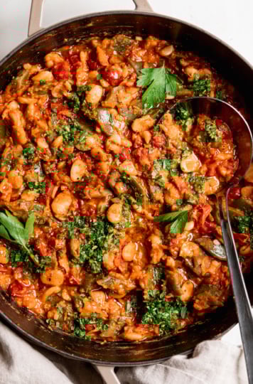 An overhead shot shows a saucy butter bean skillet with a cherry tomato sauce and parsley on top. A ladle is sticking out of the pot.