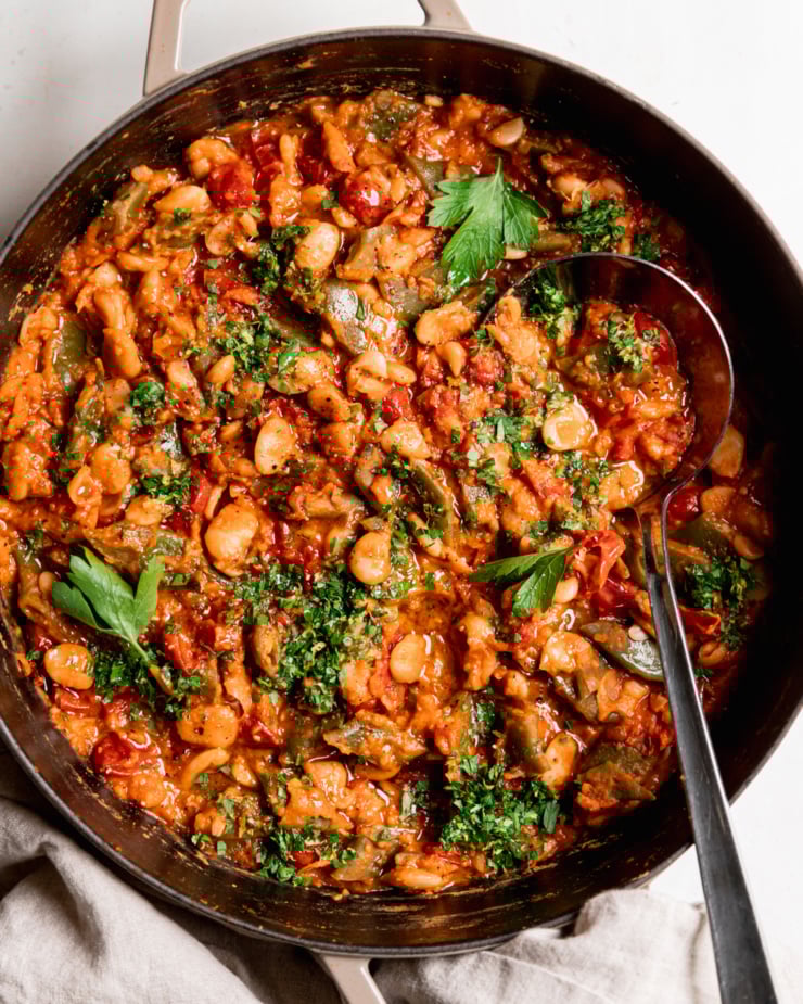 An overhead shot shows a saucy butter bean skillet with a cherry tomato sauce and parsley on top. A ladle is sticking out of the pot.