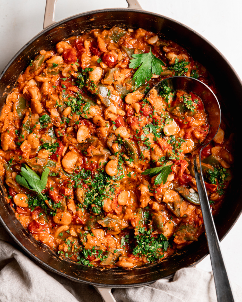 An overhead shot shows a saucy butter bean skillet with a cherry tomato sauce and parsley on top. A ladle is sticking out of the pot.