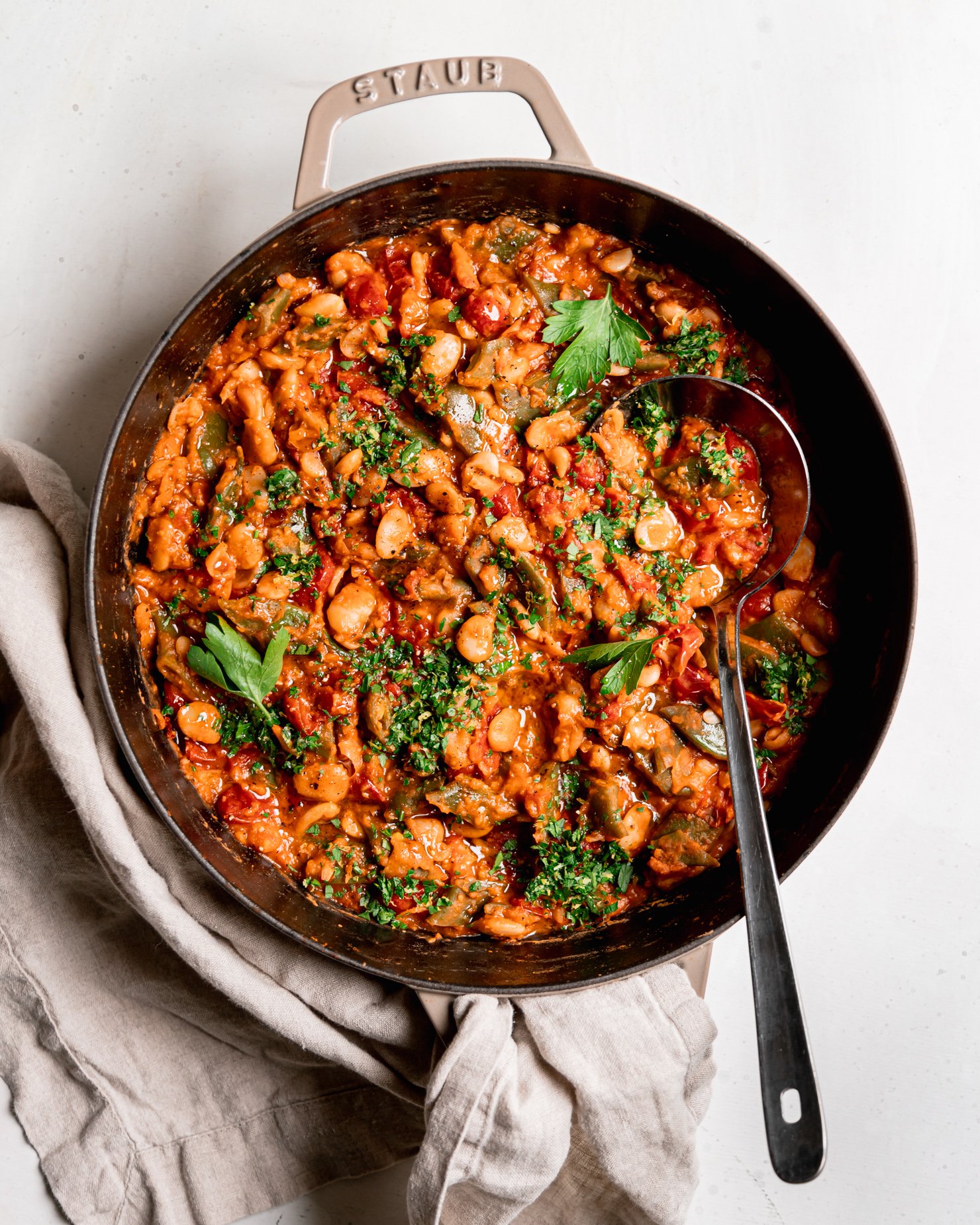An overhead shot shows a saucy butter bean skillet with a cherry tomato sauce and parsley on top. A ladle is sticking out of the pot.