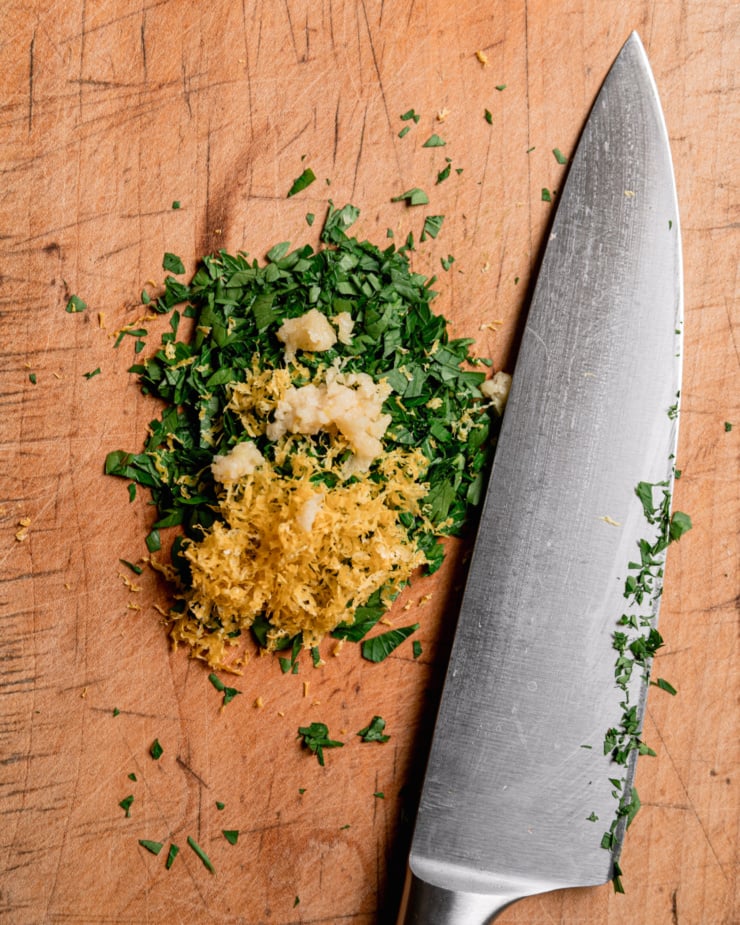An overhead shot shows gremolata (chopped parsley, lemon zest, and finely minced garlic) being made on a wooden cutting board