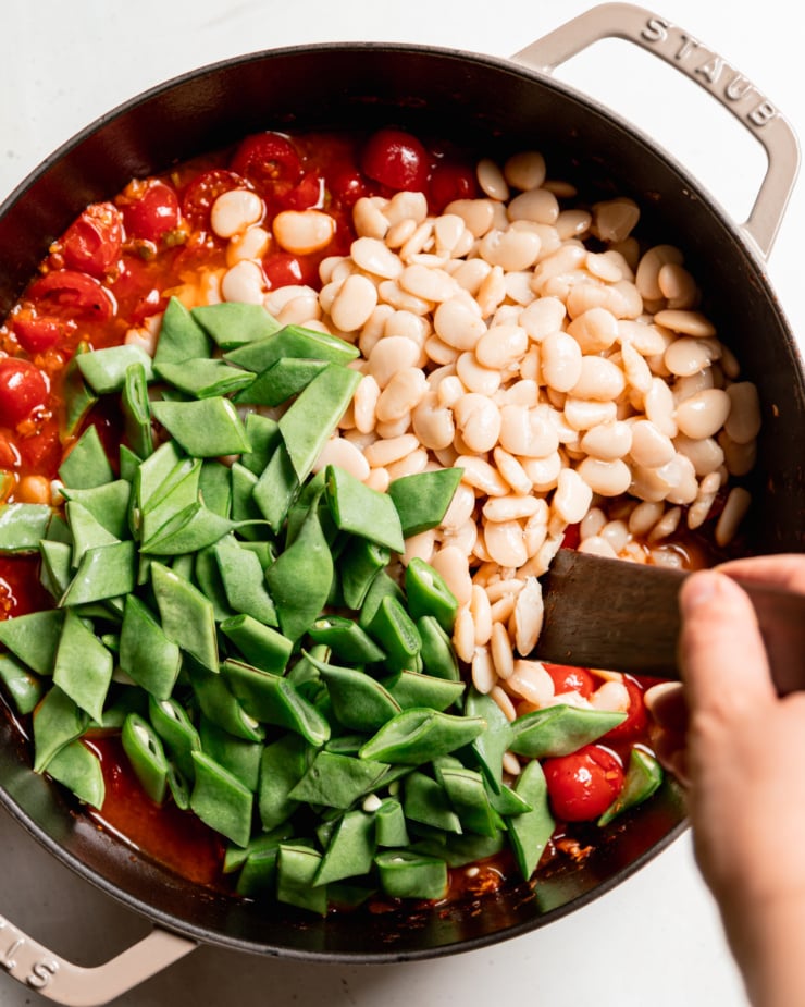 An overhead shot shows butter beans and sliced romano beans in a braser pot on top of a cherry tomato sauce mixture. A hand is using a wooden utensil to stir everything together.