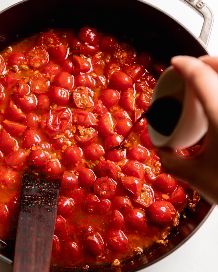 An overhead shot shows a hand pouring a small bowl of Tamari into a cherry tomato sauce mixture in a braiser-style pot.