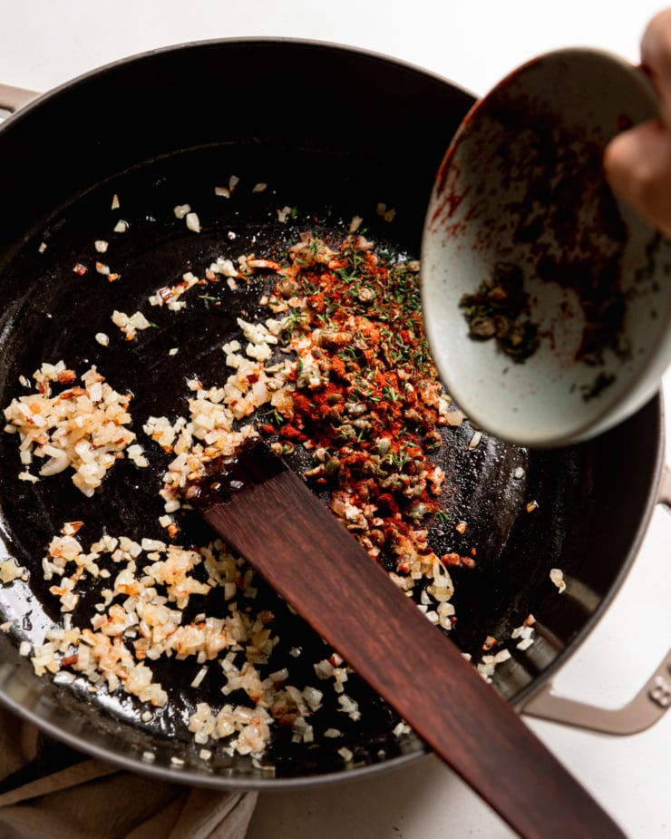 An overhead shot shows herbs, spices, and chopped capers being added to a pot with sautรฉed shallots.