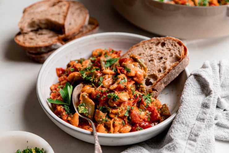 A 3/4 angle image shows a single serving of cherry tomato-sauced butter beans and green romano beans in a shallow bowl. Some whole wheat sourdough bread is sticking out of the heap of beans and a spoon is in the bowl as well.