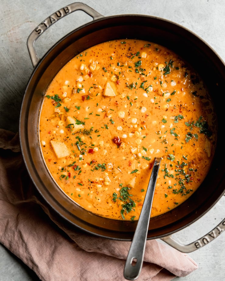 An overhead shot shows a large Dutch oven filled halfway with a smoky vegan corn chowder with potatoes. The chowder is garnished with chopped fresh herbs.