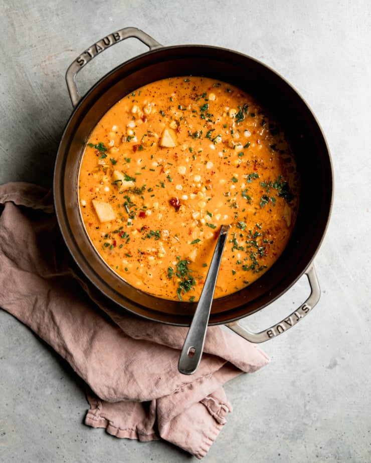 An overhead shot shows a large Dutch oven filled halfway with a smoky vegan corn chowder with potatoes. The chowder is garnished with chopped fresh herbs.