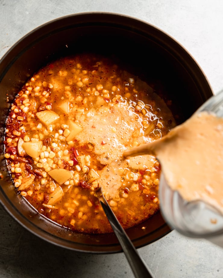 An overhead shot shows a blender pitcher emptying a blended up portion of corn chowder back into the soup pot with the remaining unblended chowder.