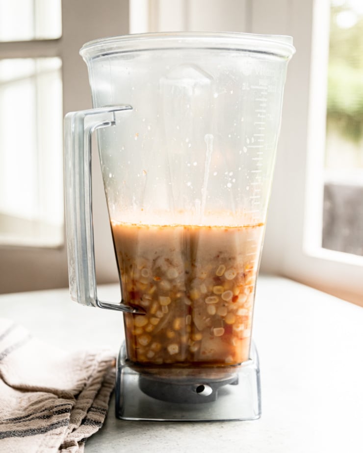 A head-on shot shows a portion of vegan corn chowder in a blender pitcher with cashew milk added.