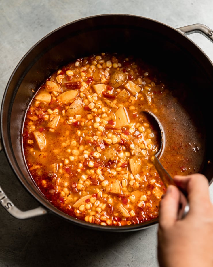 A slight 3/4 angle shot shows a hand ladling a vegan corn chowder, prior to a portion of the soup being blended.