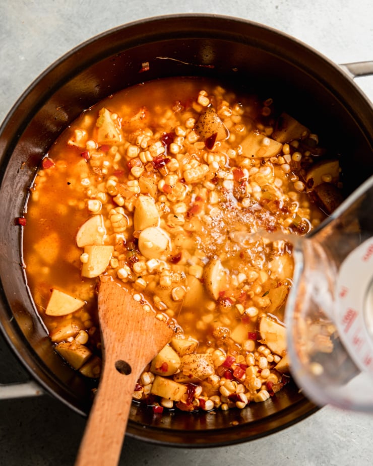 An overhead shot shows vegetable stock being added to a large pot with chopped potatoes, corn, and other vegetables.