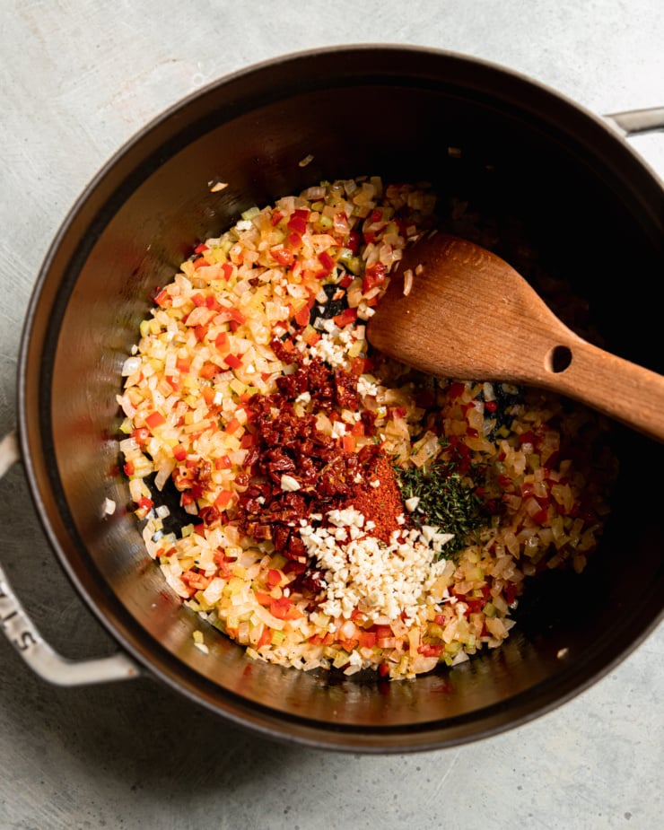 An overhead shot shows a large pot with sautéed onions, celery, and red bell pepper. Minced sun dried tomatoes, garlic, thyme, and spices have just been added to the pot. A wooden spoon is sticking out of the pot.