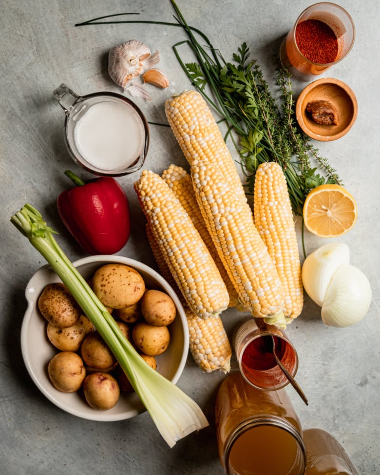 An overhead shot shows ingredients used in a vegan corn chowder recipe, including fresh cobs of corn, smoked paprika, thyme, potatoes, celery, vegetable stock and more.