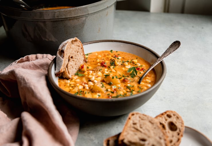 A 3/4 angle shot shows a bowl of vegan corn chowder with a piece of bread sticking out of the bowl. A soup pot is seen in the background.