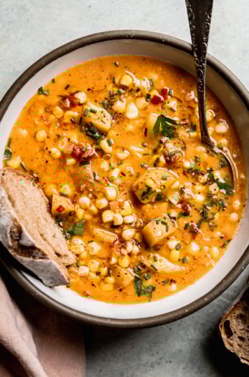 An up close, overhead shot shows a single serving of vegan corn chowder with potatoes in a ceramic bowl. A piece of whole wheat sourdough is sticking out of the bowl. The chowder is garnished with chopped fresh herbs and ground black pepper.