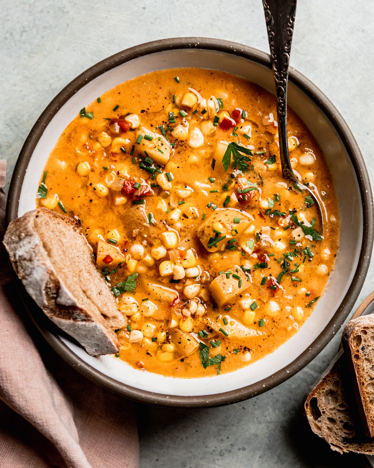An up close, overhead shot shows a single serving of vegan corn chowder with potatoes in a ceramic bowl. A piece of whole wheat sourdough is sticking out of the bowl. The chowder is garnished with chopped fresh herbs and ground black pepper.