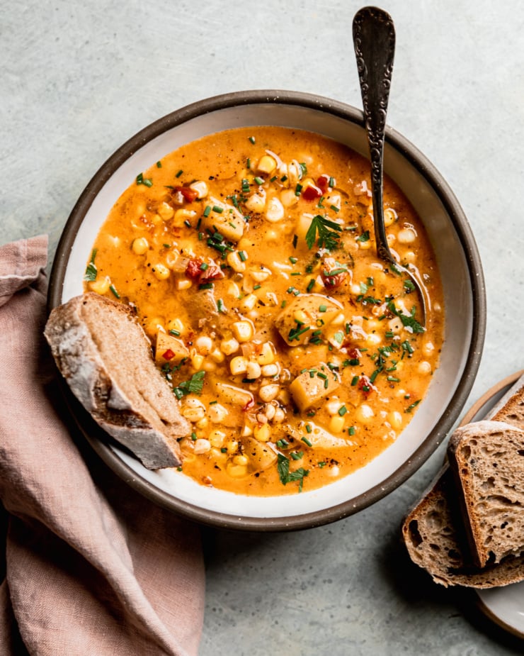 An overhead shot shows a single serving of vegan corn chowder with potatoes in a ceramic bowl. A piece of whole wheat sourdough is sticking out of the bowl. The chowder is garnished with chopped fresh herbs and ground black pepper.