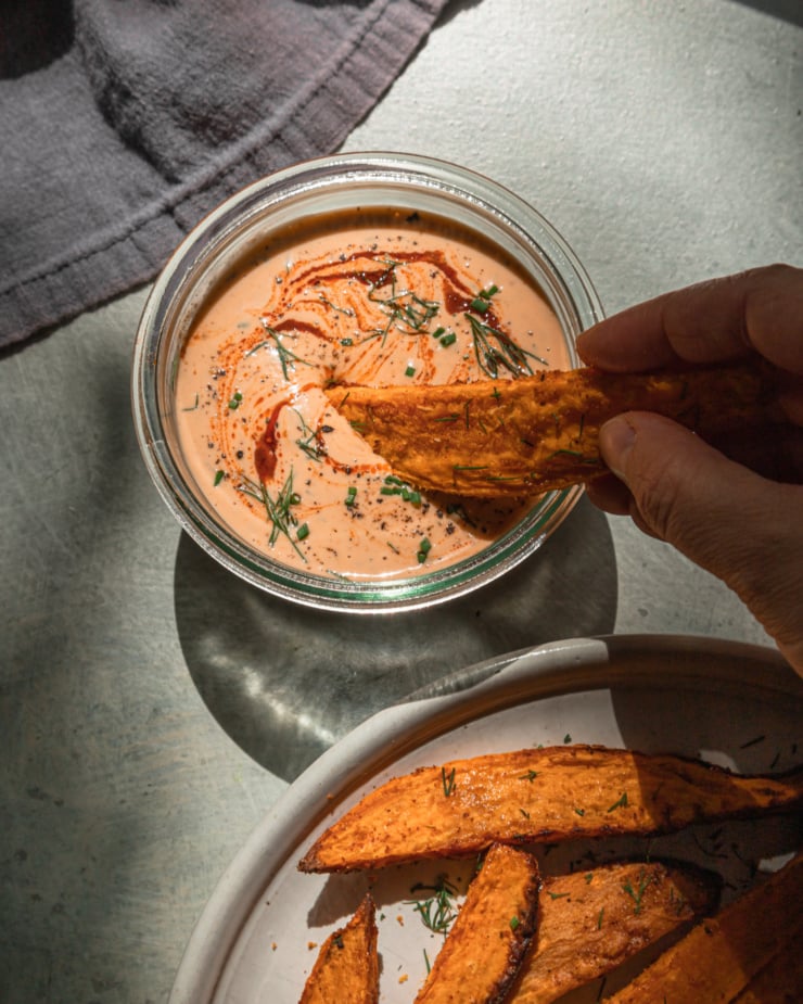 An overhead shot shows a hand dipping a roasted sweet potato wedge into a container of creamy spicy tahini ranch sauce.