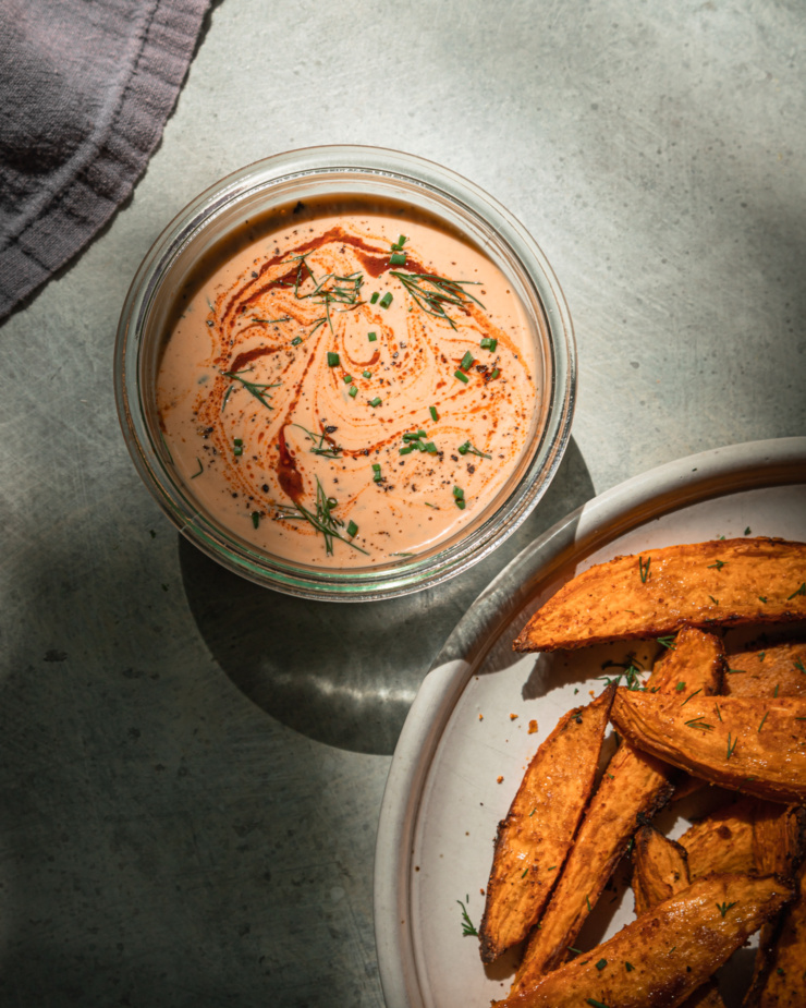 An overhead shot shows a round container of creamy sauce with some baked sweet potato wedges on a plate nearby.