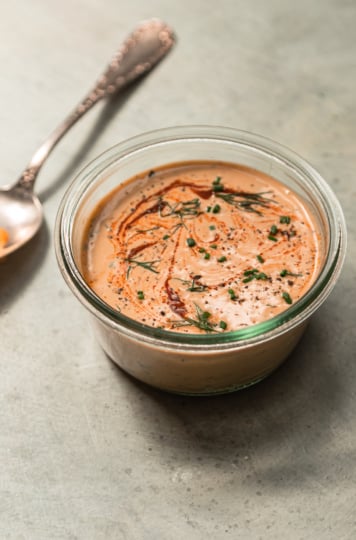 A 3/4 angle shot shows a container of spicy tahini ranch in bright natural light. The top is swirled with hot sauce and garnished with chopped herbs.