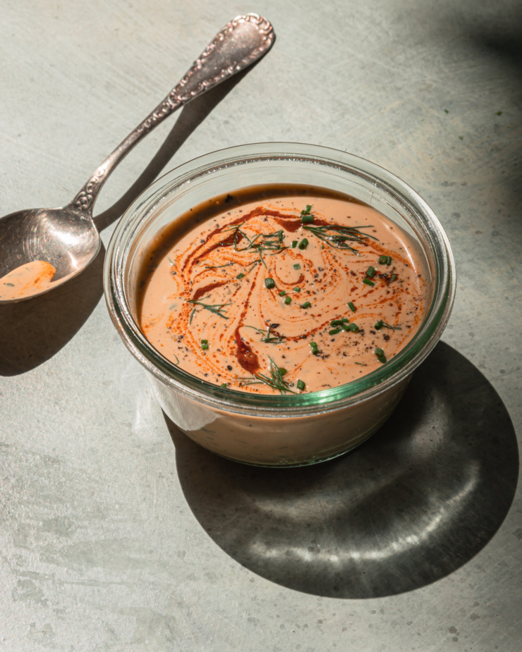 A 3/4 angle shot shows a container of spicy tahini ranch in direct sunlight. The top is swirled with hot sauce and garnished with chopped herbs.