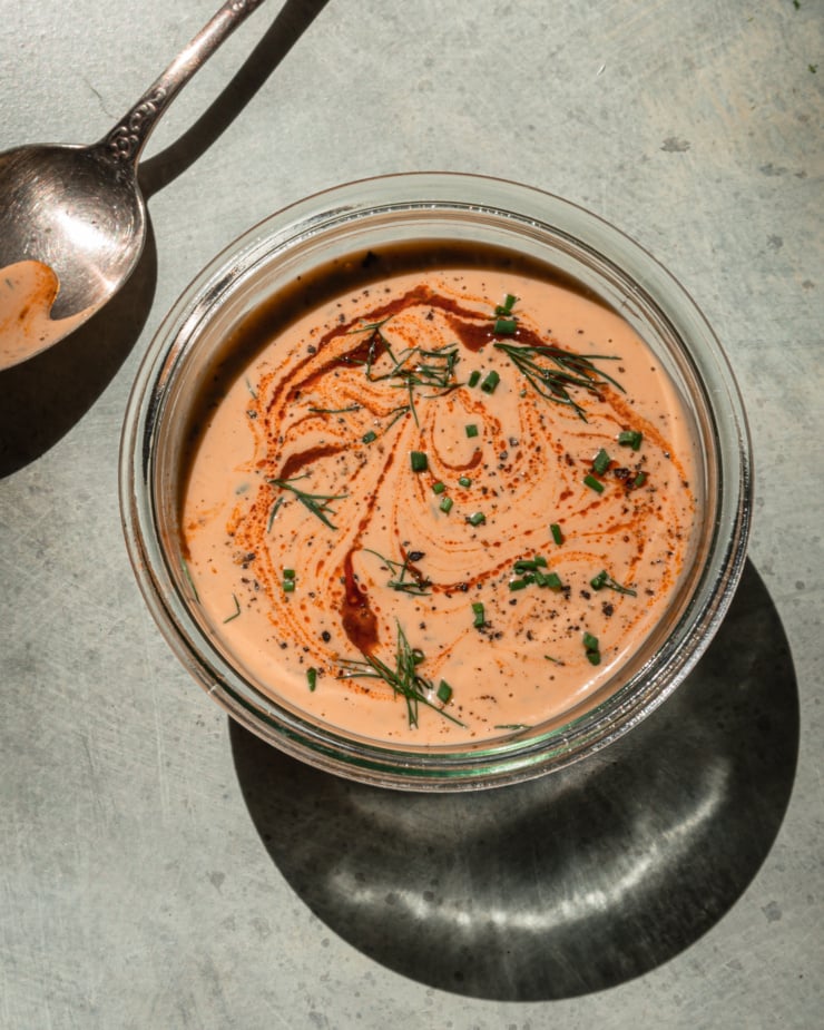 An overhead shot shows a container of spicy tahini ranch in direct sunlight. The top is swirled with hot sauce and garnished with chopped herbs.