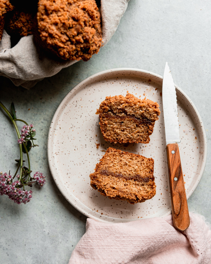 An overhead shot shows a vegan coffee cake muffin split in half on top of a speckled ceramic plate. A wood handled knife is perched in the plate as well as a flower from a sedum plant.