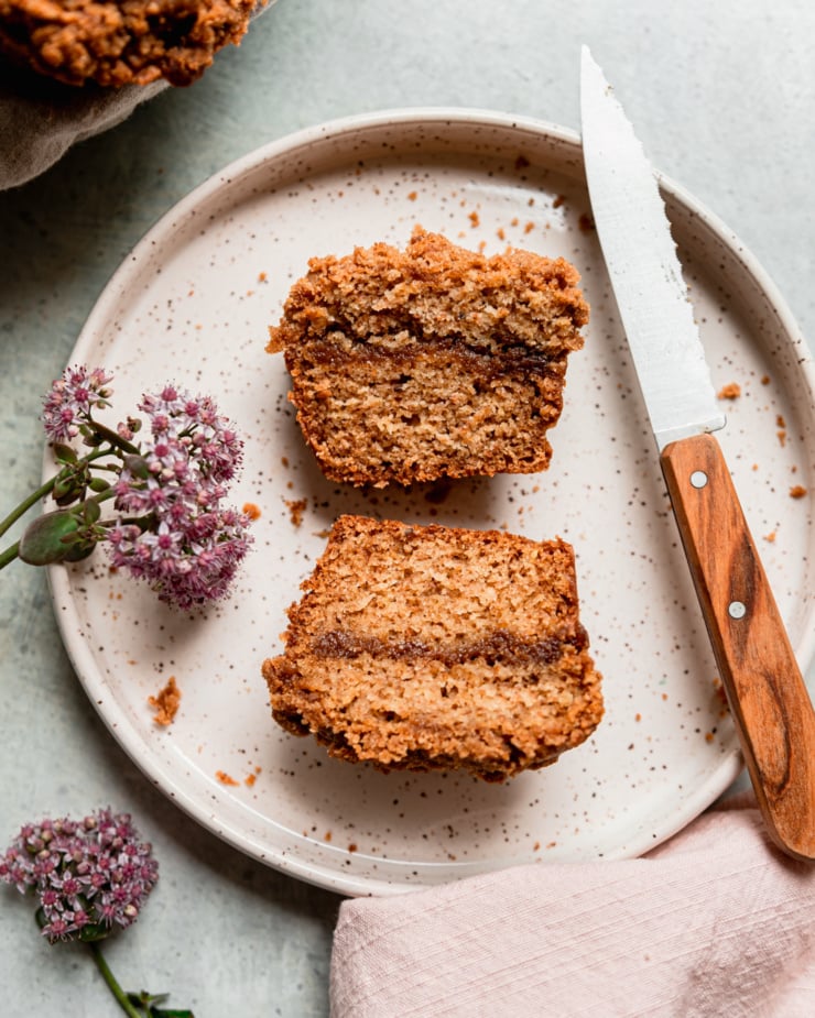 An overhead shot shows a vegan coffee cake muffin split in half on top of a speckled ceramic plate. A wood handled knife is perched in the plate as well as a flower from a sedum plant.