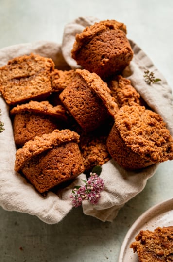 A 3/4 angle shot shows vegan coffee cake muffins arranged in a linen lined bowl with some sedum flowers placed nearby.