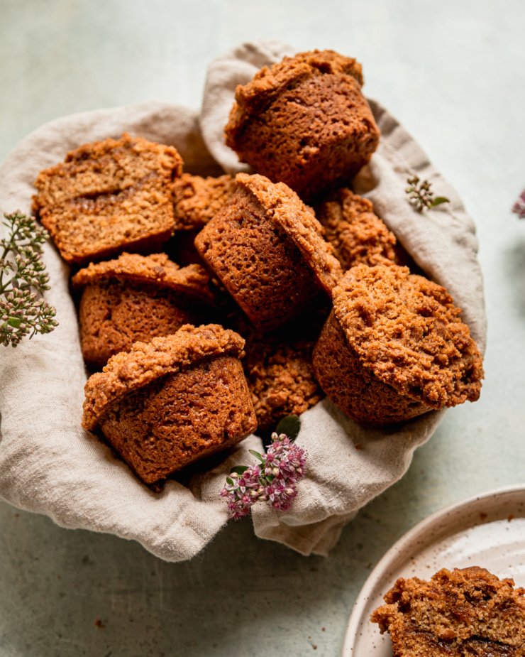 A 3/4 angle shot shows vegan coffee cake muffins arranged in a linen lined bowl with some sedum flowers placed nearby.