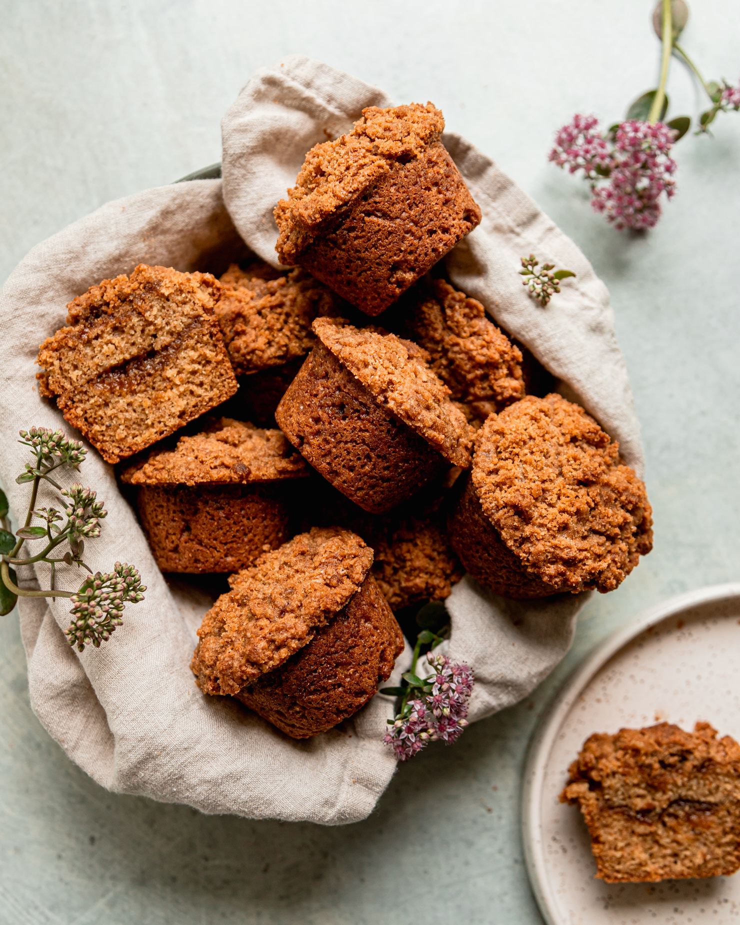 An overhead shot shows vegan coffee cake muffins arranged in a linen lined bowl with some sedum flowers placed nearby.