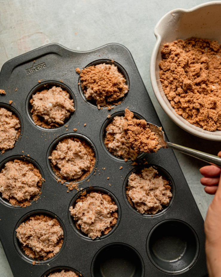 An overhead shot shows a hand using a spoon to portion streusel topping on top of portions of muffin batter.