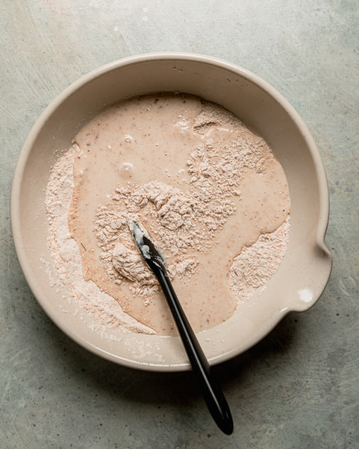 An overhead shot shows liquid ingredients that were recently added to a flour mixture in a ceramic mixing bowl. A spatula is sticking out of the bowl.
