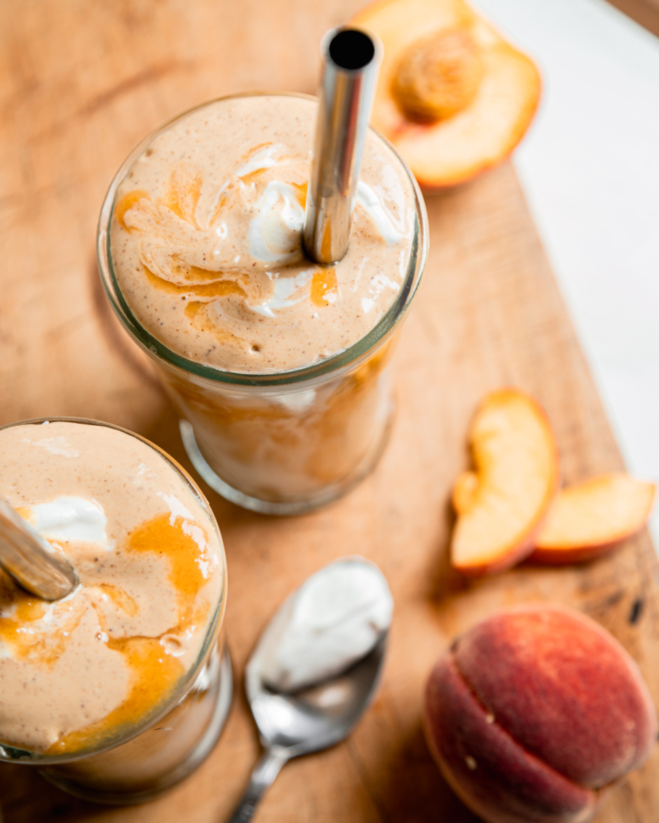 An overhead shot of two glasses of peaches and cream protein smoothie. A spoon of yogurt, a whole peach, and peach slices are seen nearby. Metal straws are sticking out of the smoothies.